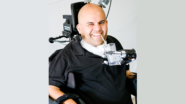 Photo of Erick Sorto drinking a glass of water assisted by a robotic arm that he controls with his thoughts. 