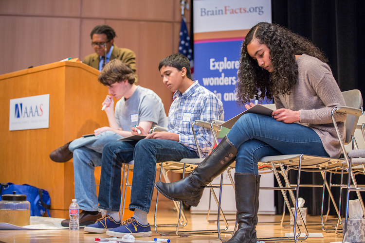 Three high school students compete in the final round of the DC Brain Bee while moderator Ben Walker looks on.