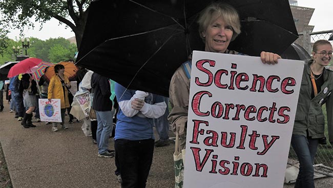 Marcher stands with a black umbrella with a sign that reads: "Science Corrects Faulty Vision."
