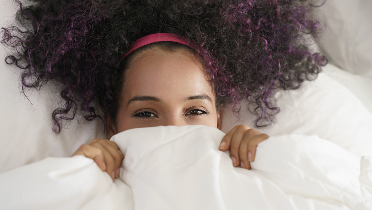 Photograph of a smiling woman in bed with a blanket covering her mouth